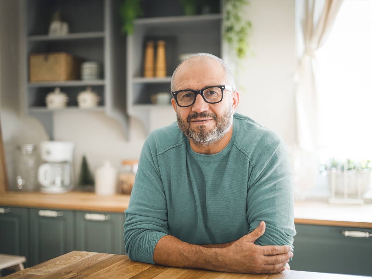 Older white man wearing glasses and a light blue shirt leans on the countertop in the kitchen