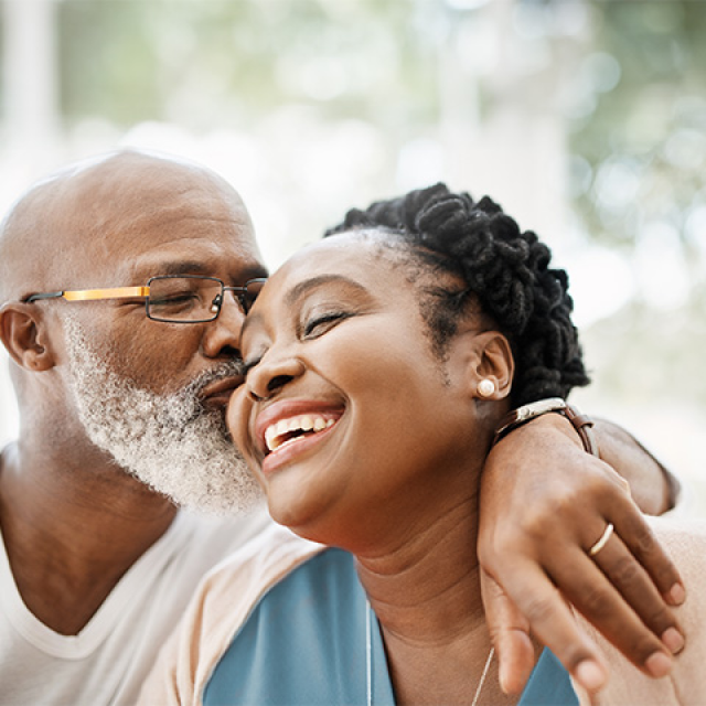 African American man with grey beard and glasses kisses a smiling African American woman on the cheek