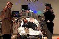 Three men in costume engage in simulation activities around a hospital bed, showcasing a medical training scenario.