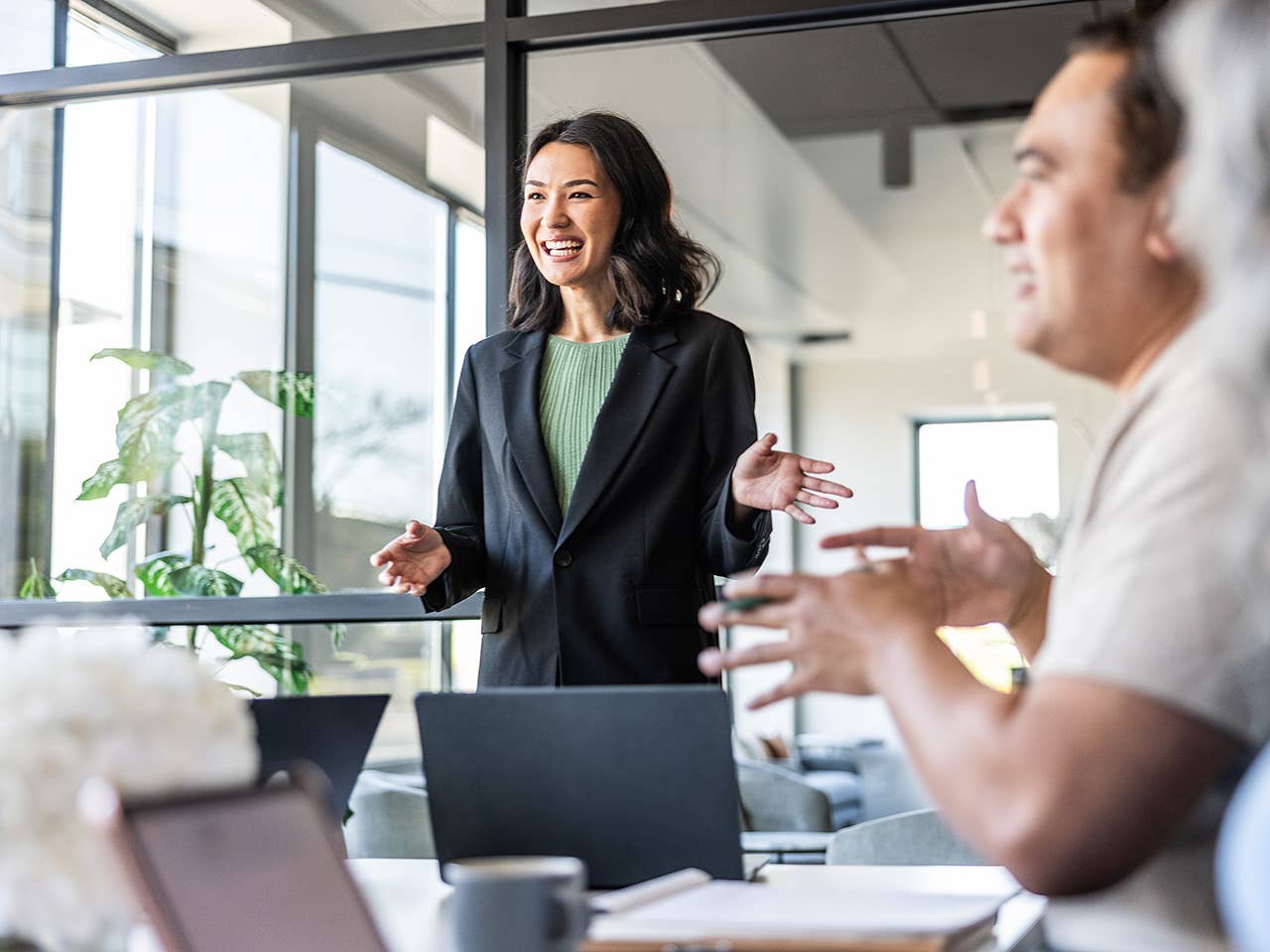 Smiling Asian woman wearing a green blouse and black sport coat stands and leads a discussion in a board room meeting