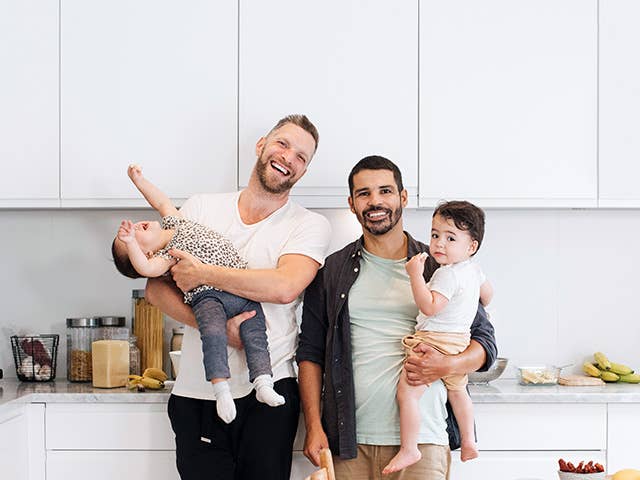 Two smiling men stand side by side as they lift up and hold their young children