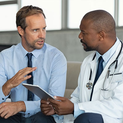 White male in a shirt and tie speaks with Black male in a lab coat