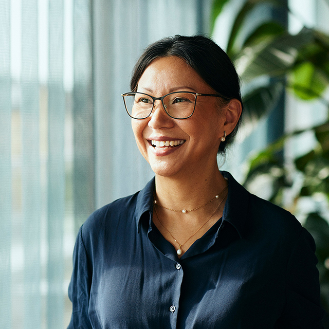 A smiling woman wearing glasses stands in front of a window, with natural light illuminating her face.