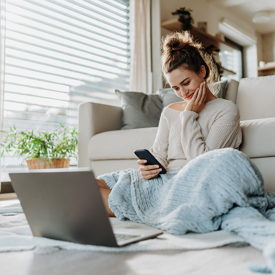 Young woman sits on living room floor with a blanket, looking at her phone