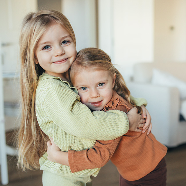 Two young girls with long blonde hair smile and hug each other in a living room