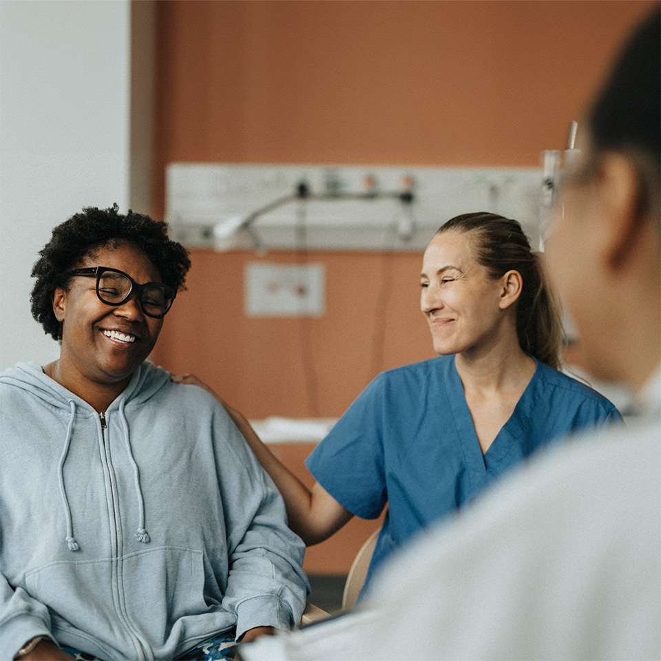 Smiling medical worker in blue scrubs places hand on shoulder of smiling Black woman sitting next to her