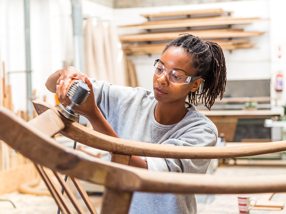 Young Black woman uses a power sander on curved wood in a shop