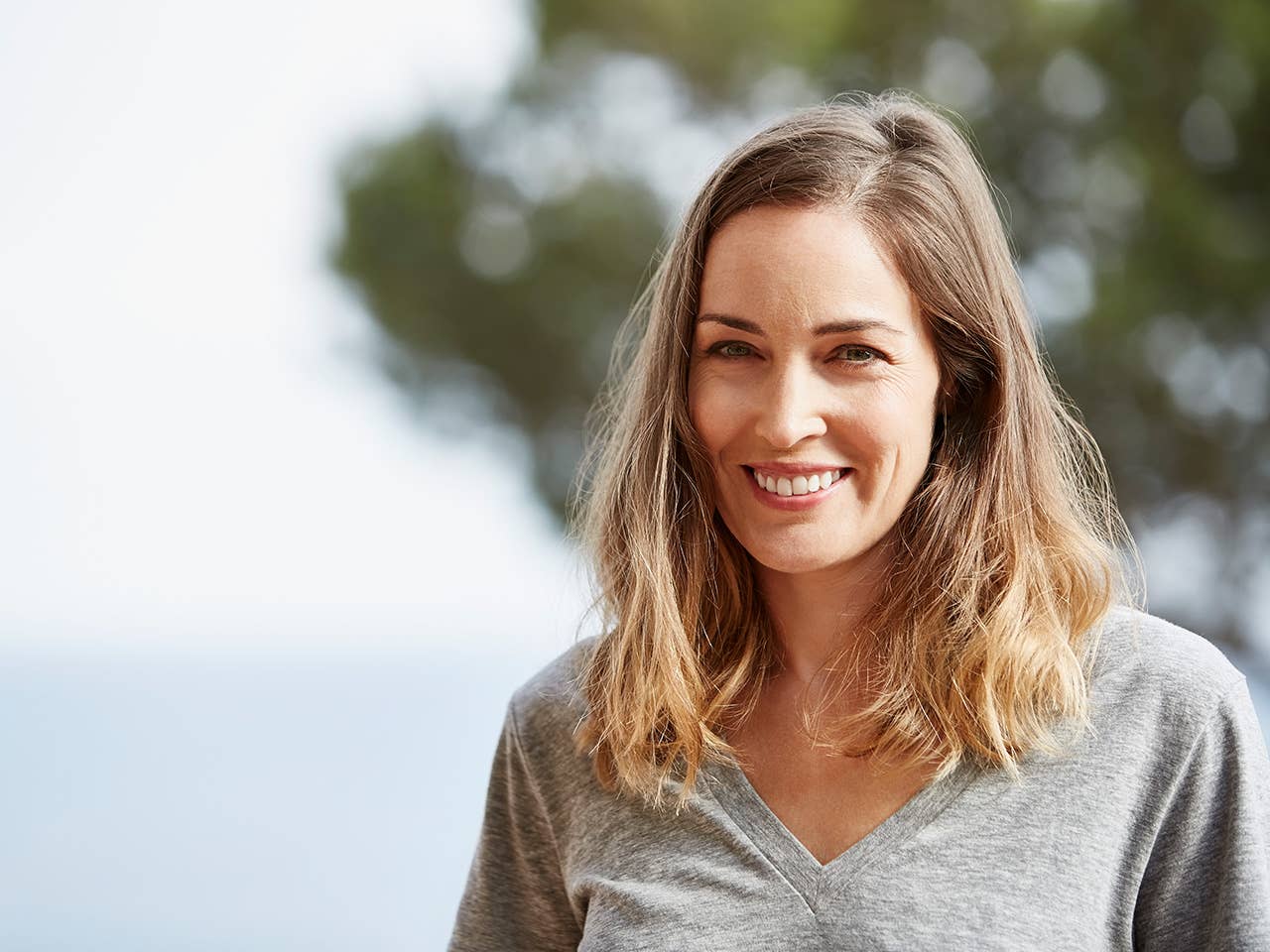 Smiling White woman in a grey shirt stands outdoors in front of a tree