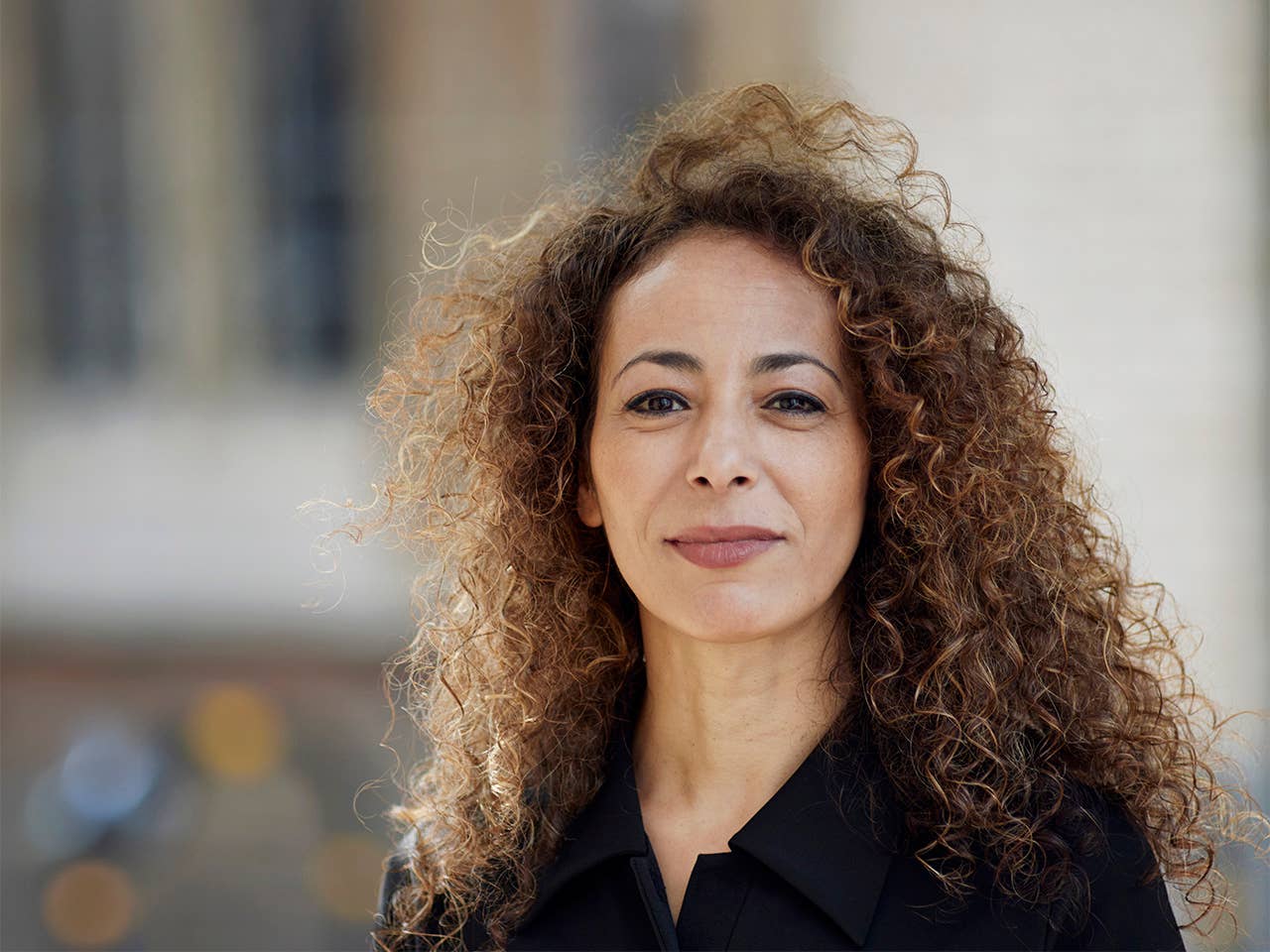 White woman with curly brown hair stands outdoors in front of a building
