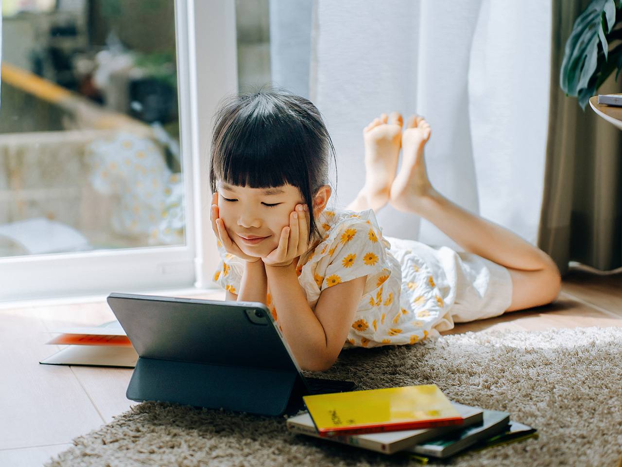 An Asian girl wearing a white shirt with yellow polka dots lies on the floor in a sunlit room looking at a tablet device