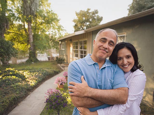 Man and his wife stand next to one another outdoors in the evening next to their home