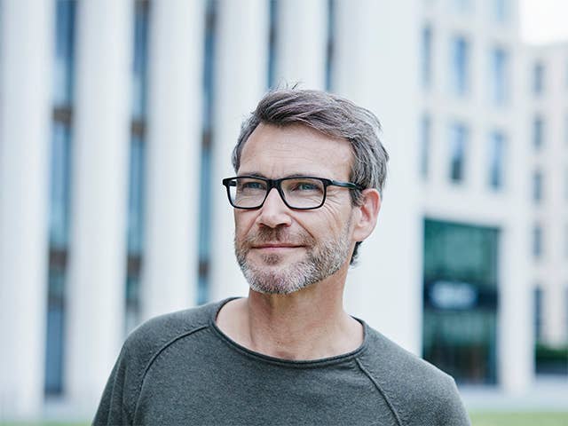 Middle aged white man with glasses and a grey beard stands outdoors in front of an office building