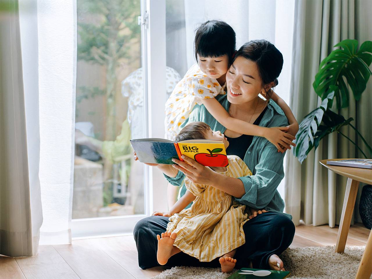 Smiling young Asian woman sits in the sunlight reading a book to her young daughter and son