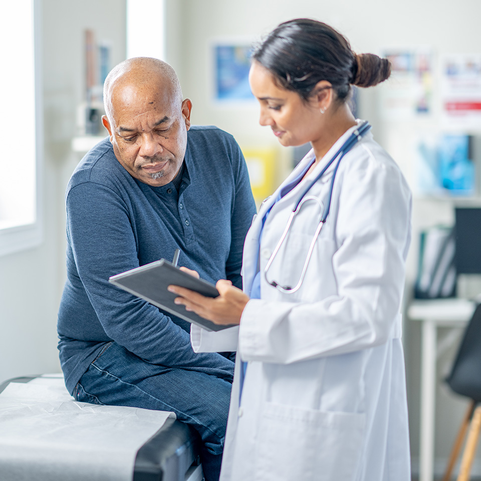 Black man looks at tablet being held by female doctor as they talk in an exam room