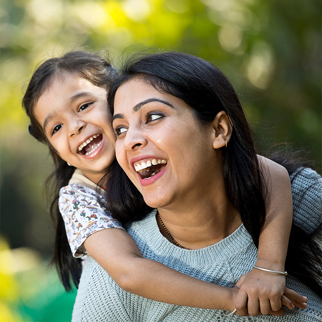 Woman with long black hair laughs while carrying her smiling daughter on her back outdoors
