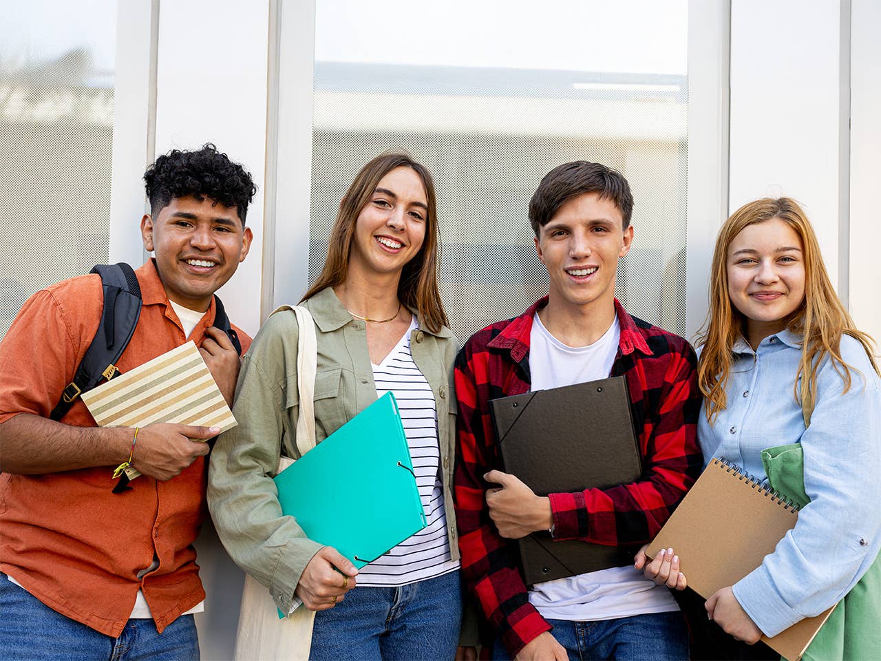 Four students stand together, smiling, each holding a stack of books in a bright, outdoor setting.