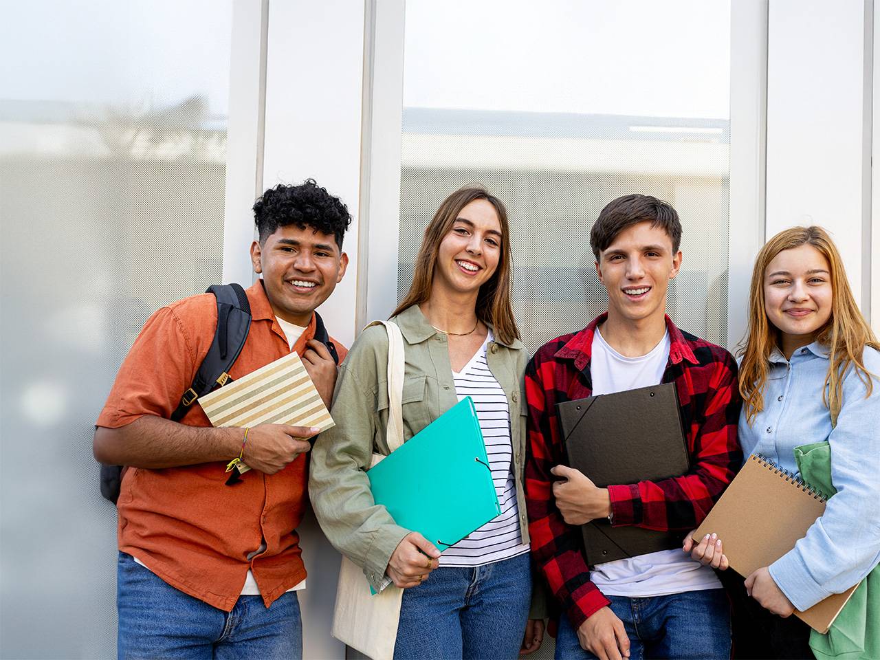 Four students stand together, smiling, each holding a stack of books in a bright, outdoor setting.