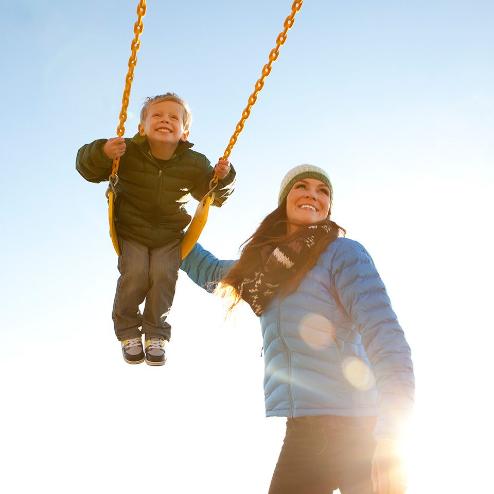 Smiling mother in a winter coat and hat pushes her smiling son on a swing