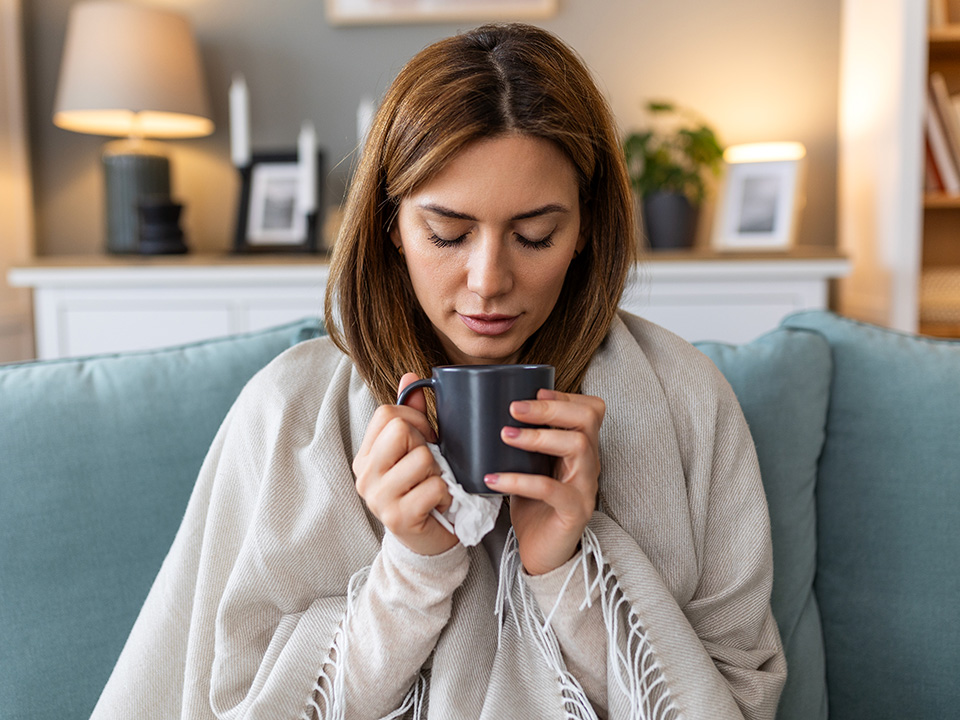 Person wrapped in a blanket holding a warm drink while recovering from flu symptoms at home.