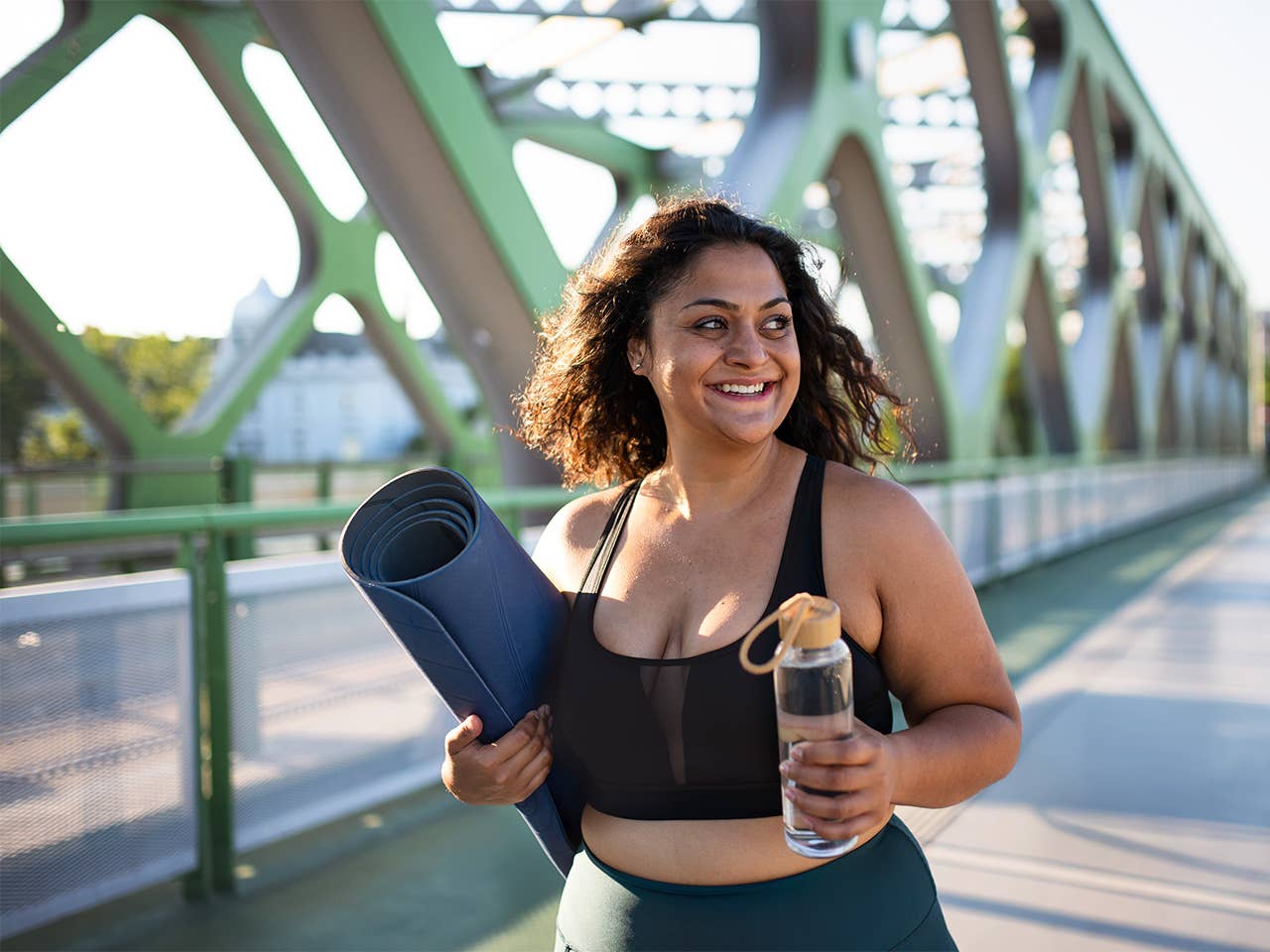 Middle aged woman carries a yoga mat and a water bottle outdoors in the sunshine