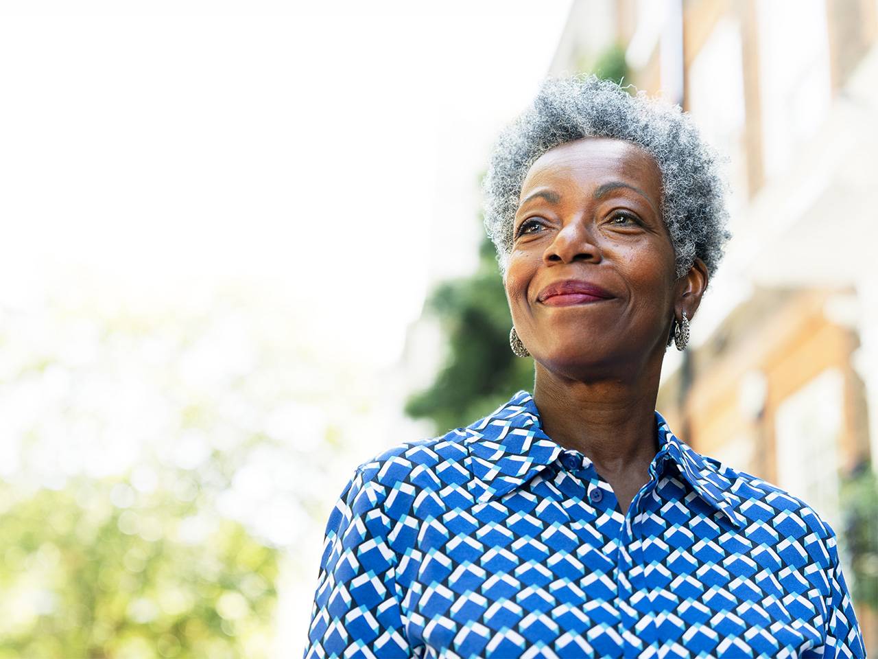 Smiling Black woman with short grey hair stands in the shade outdoors in front of a brick building
