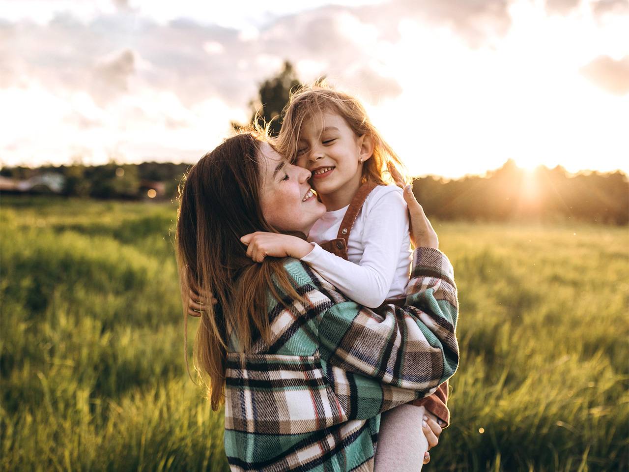 Smiling woman wearing a plaid shirt lifts and hugs her smiling daughter outdoors in the sunshine