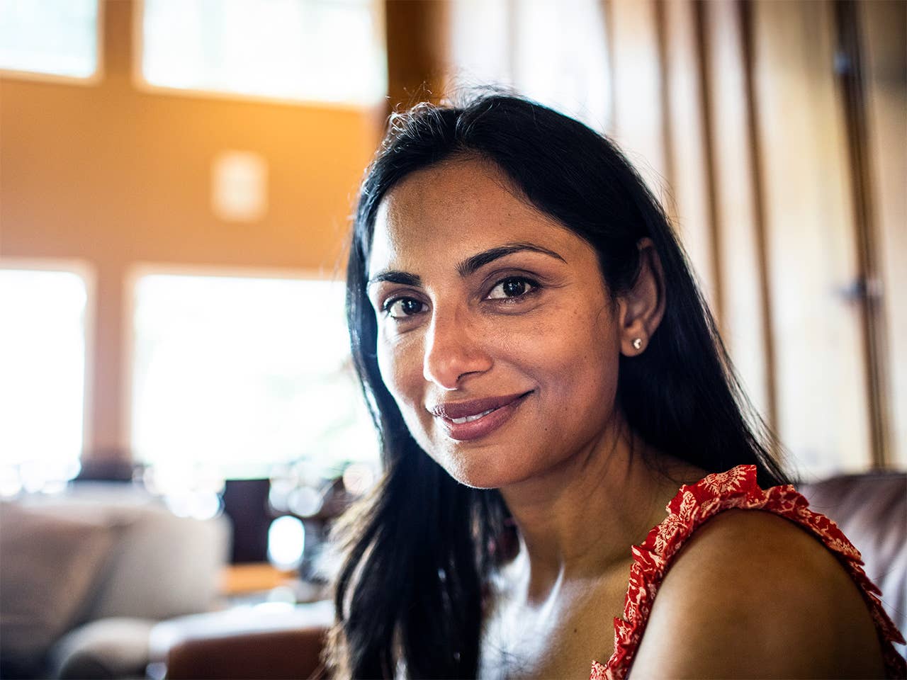 Smiling woman with long black hair sits indoors near a sunshine filled window