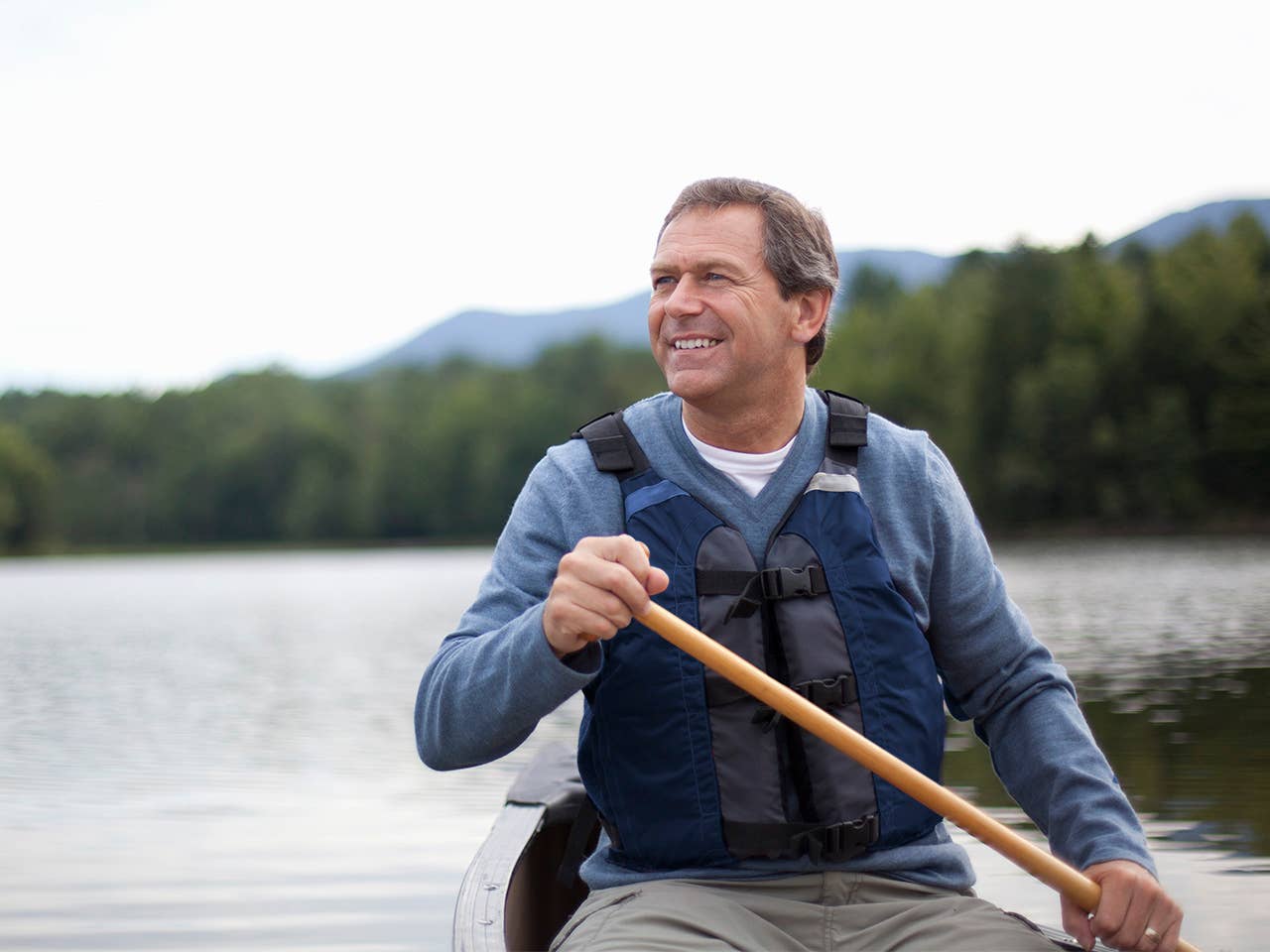 Older white man wearing a navy blue life jacket and holding an oar sits in a boat on a lake