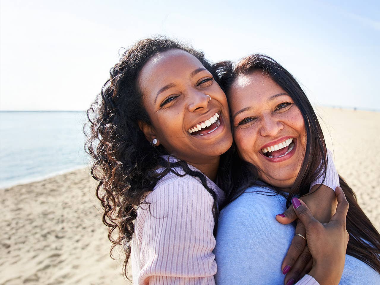 Smiling young woman with curly brown hair hugs her smiling mother while they stand in the sunshine on a beach