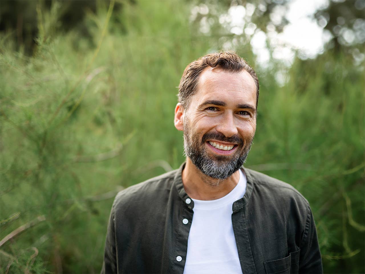 Smiling man with brown hair and a beard standing in outdoors in front of trees