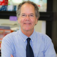 A man in a blue shirt and tie stands in front of a lab, representing Dr. Charles E. Schwartz, a research director.