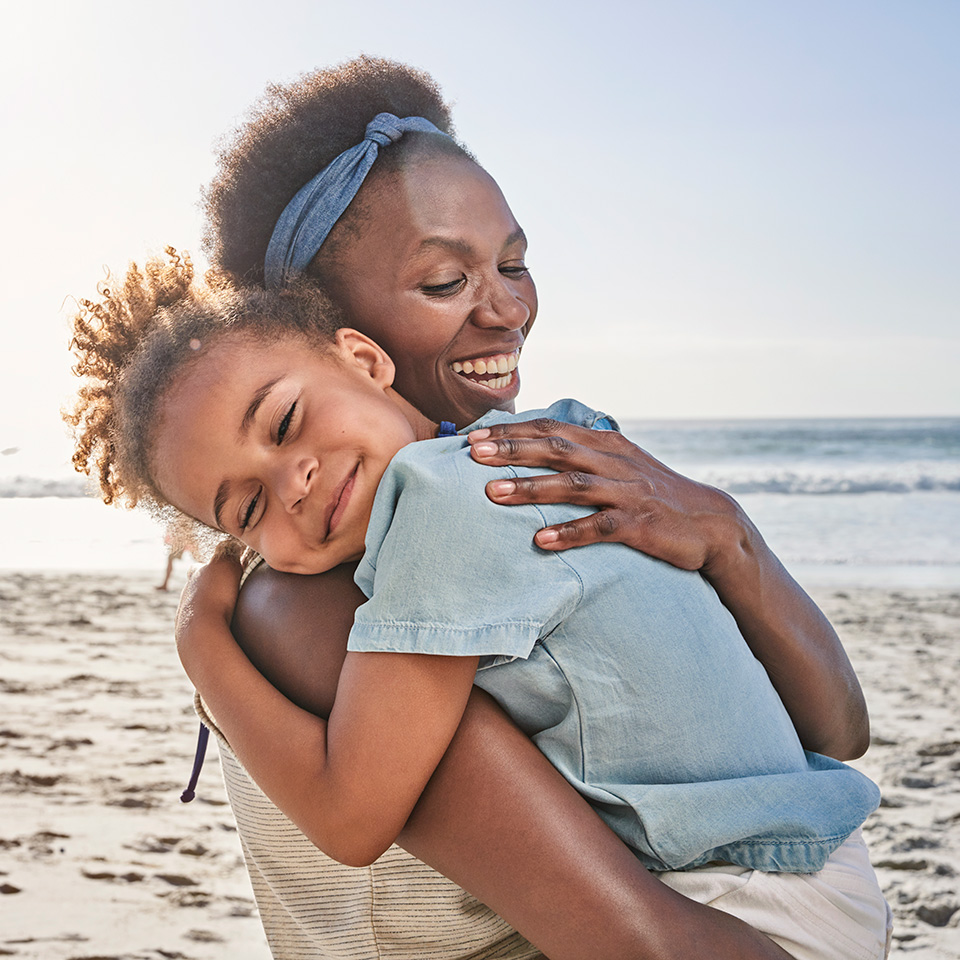 African American woman holds and hugs young African American girl while standing on beach in the sunshine