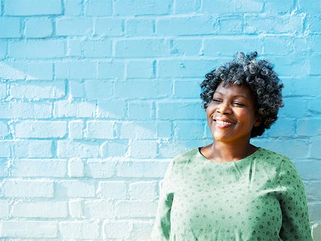 Smiling middle aged Black woman wearing a light green blouse stands outside in front of a white brick wall