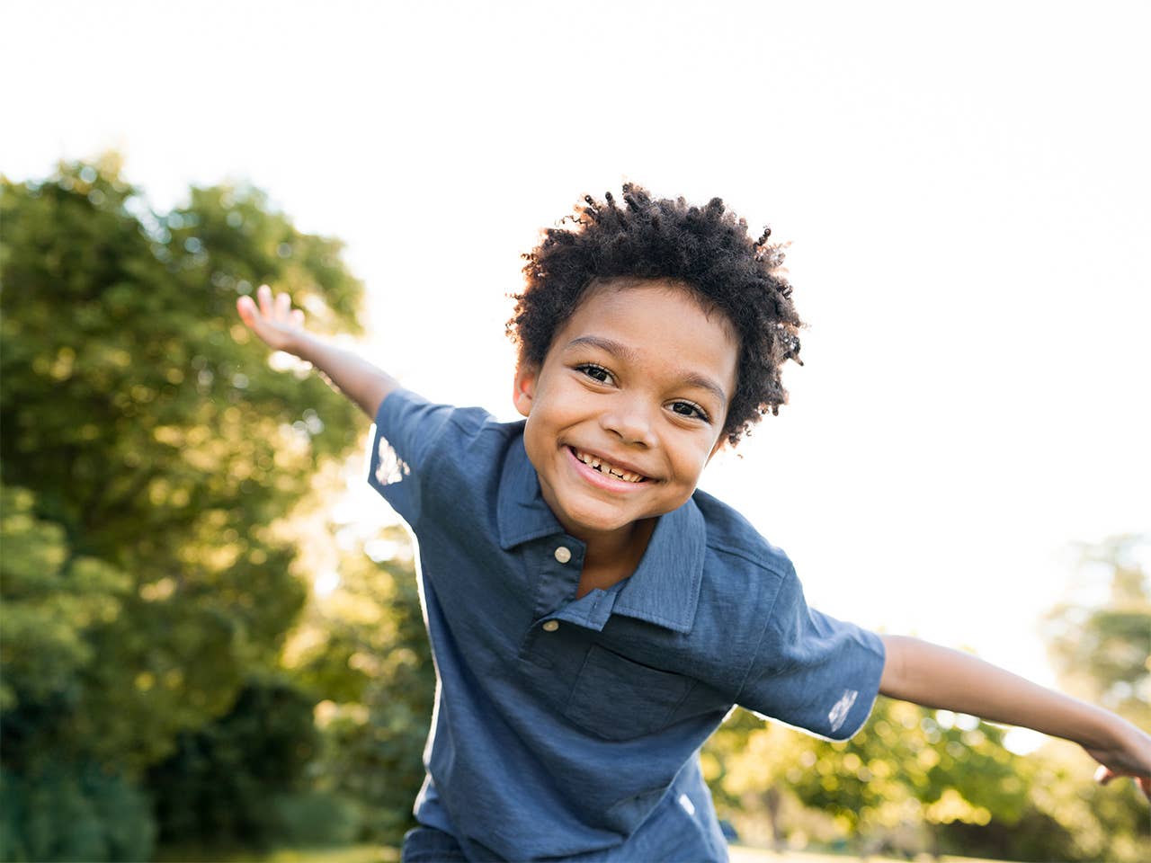 Smiling Black boy with curly brown hair leans forward in the sunshine with his arms spread out like wings
