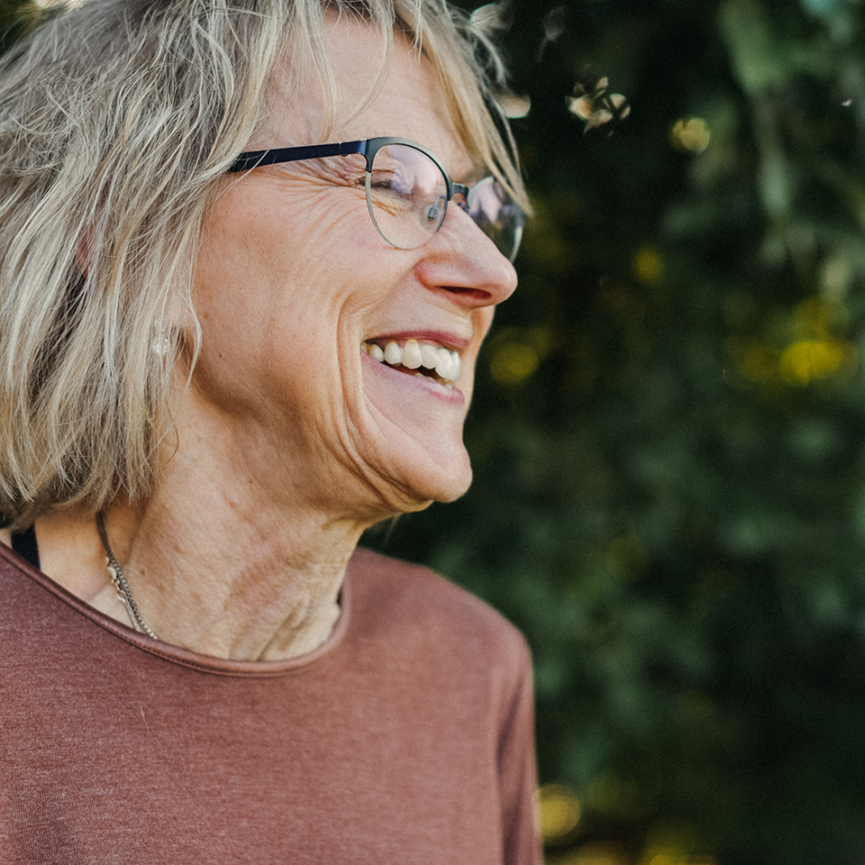 Smiling older woman with grey hair and glasses stands in front of trees