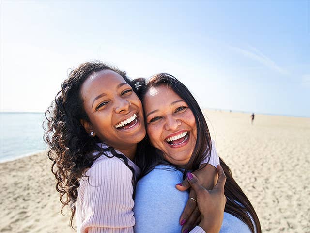 Smiling young woman with curly brown hair hugs her smiling mother while they stand in the sunshine on a beach