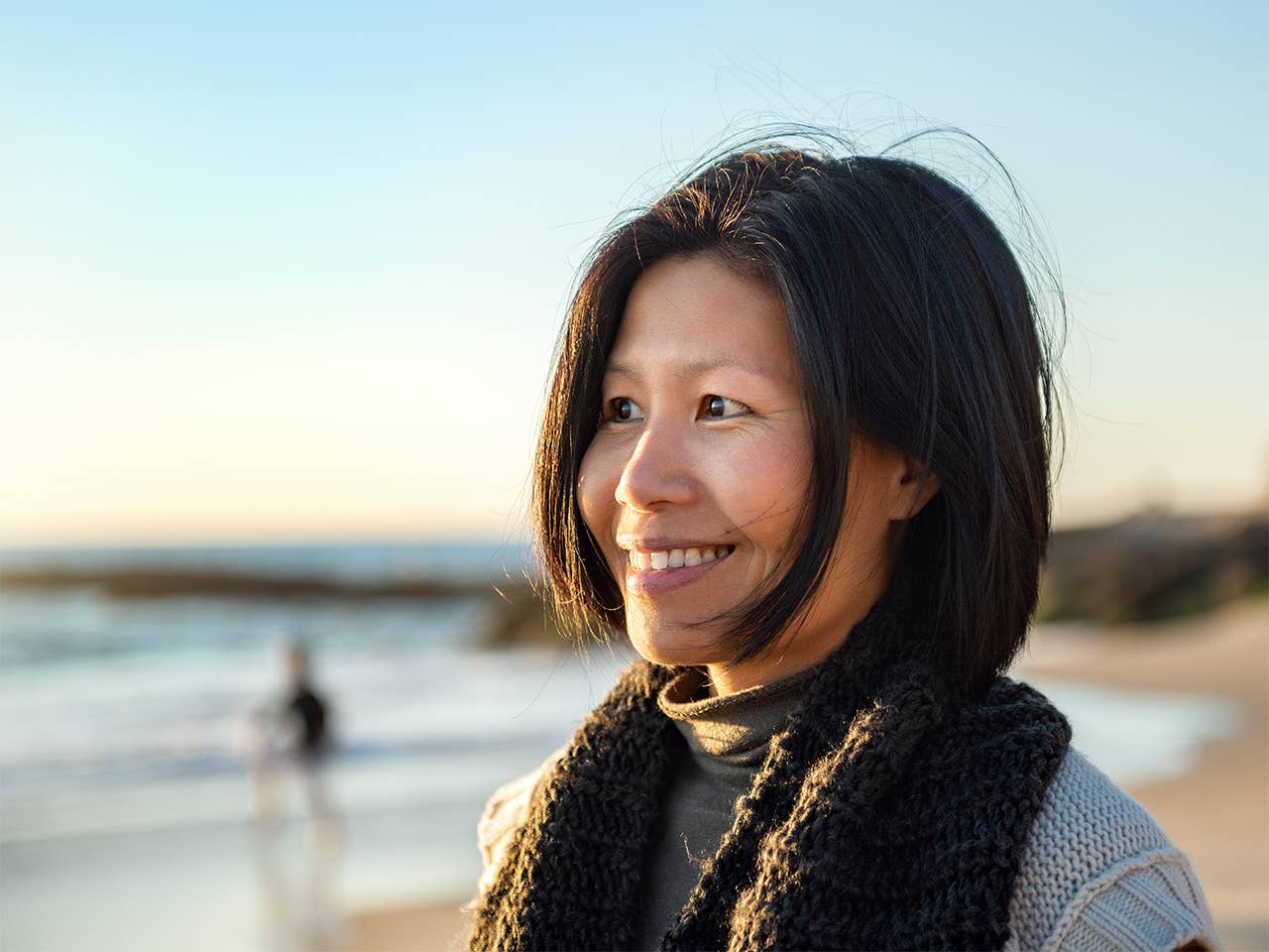 A woman smiles brightly while standing on a sandy beach with gentle waves in the background.