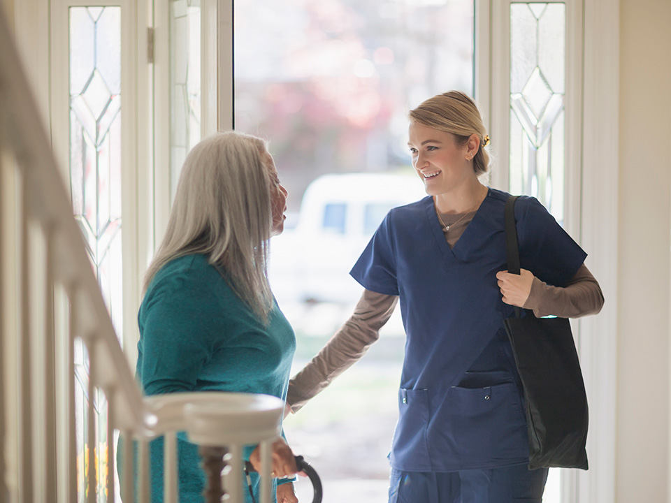 An at-home care nurse wearing navy blue scrubs smiles and speaks with a woman in her house