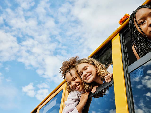 Three smiling grade-school age girls stick their heads out from school bus windows