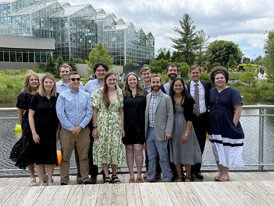 Group of 12 medical residency trainees posing on a boardwalk beside a pond, with a large glass conservatory behind them.