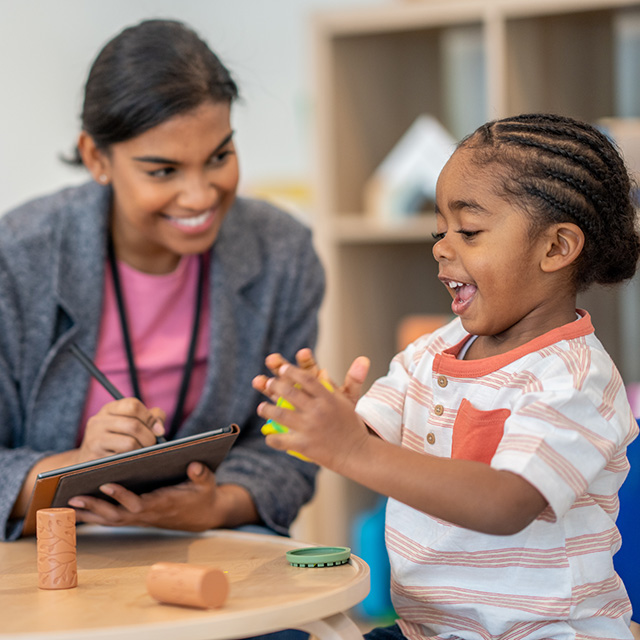 Smiling Corewell Health Child Life Specialist sits at a table next to a laughing young Black girl who is playing with clay