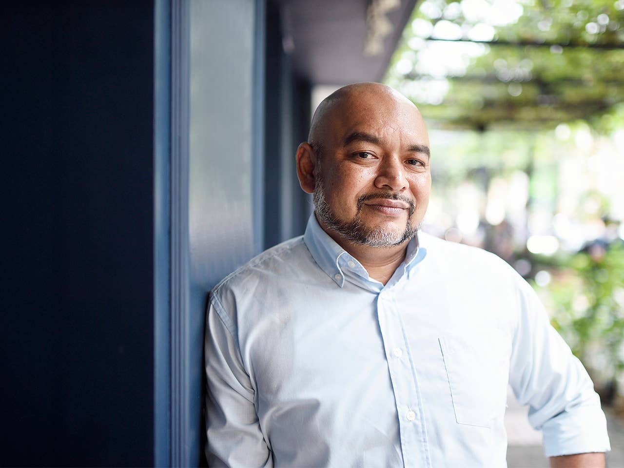 Man with a grey goatee and bald head leans against a blue building while standing outdoors