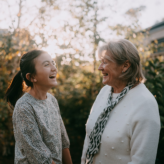 A teenage girl with black hair in a ponytail laughs while speaking to her grandmother outdoors as the sun sets behind trees