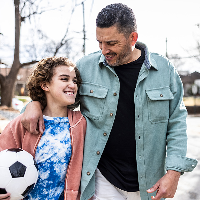 A smiling girl wearing a blue and white shirt looks at her dad as they hug outdoors in the sunshine