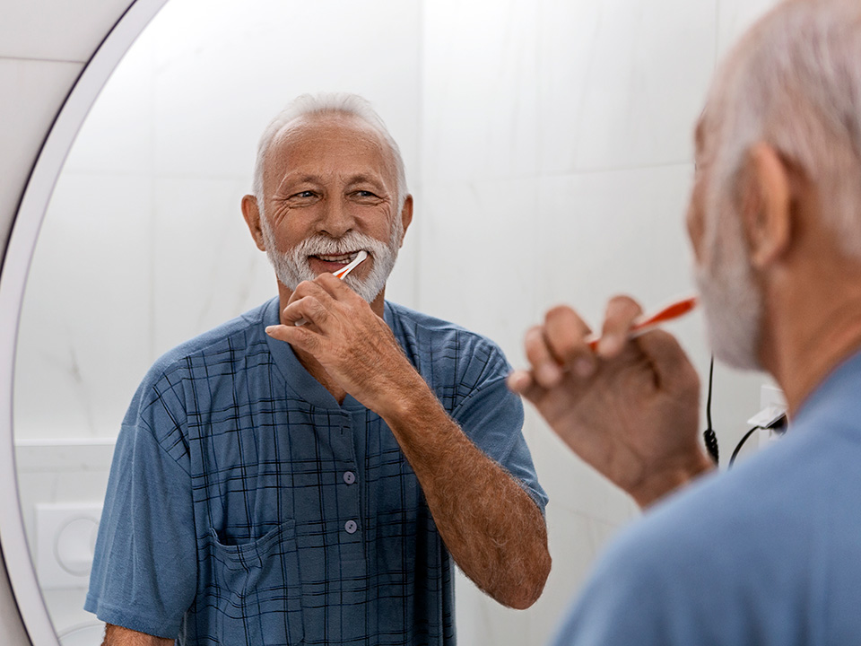 Elderly man in a blue shirt looks in the mirror while brushing his teeth