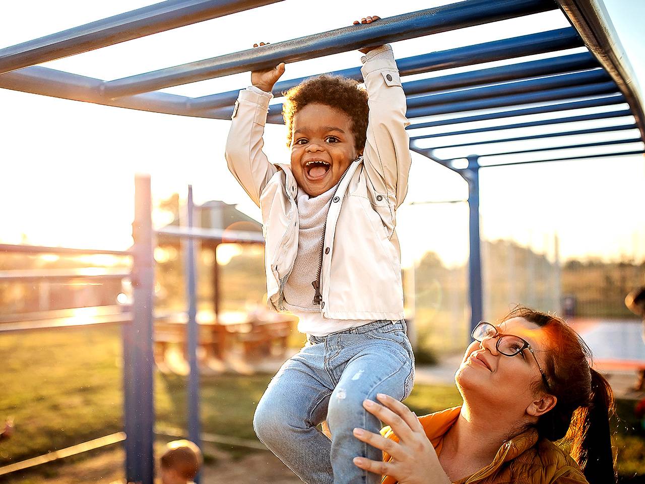 Smiling young Black boy wearing jeans and a white jacket hangs from monkey bars on a playground