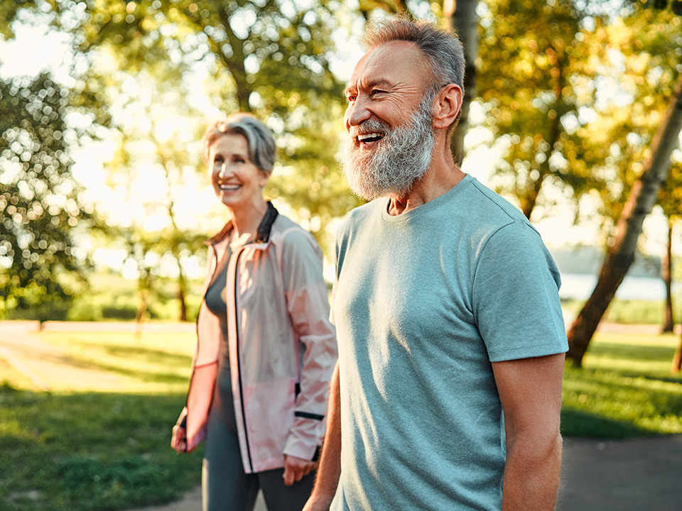 Older white man and woman smile while walking outdoors in the sunshine