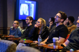 A group of people seated in a seminar, observing a presentation on a screen, with some taking notes.