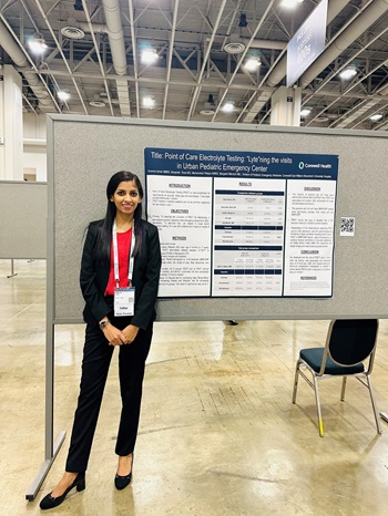 A woman stands beside a conference poster, engaging with attendees about pediatric emergency medicine research topics.