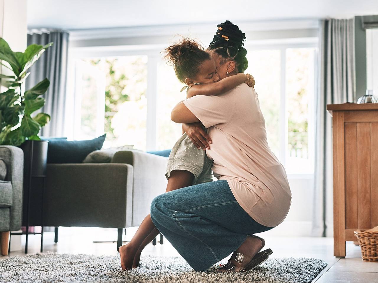 A Black woman kneels down and hugs her young daughter while they are in a sunny living room